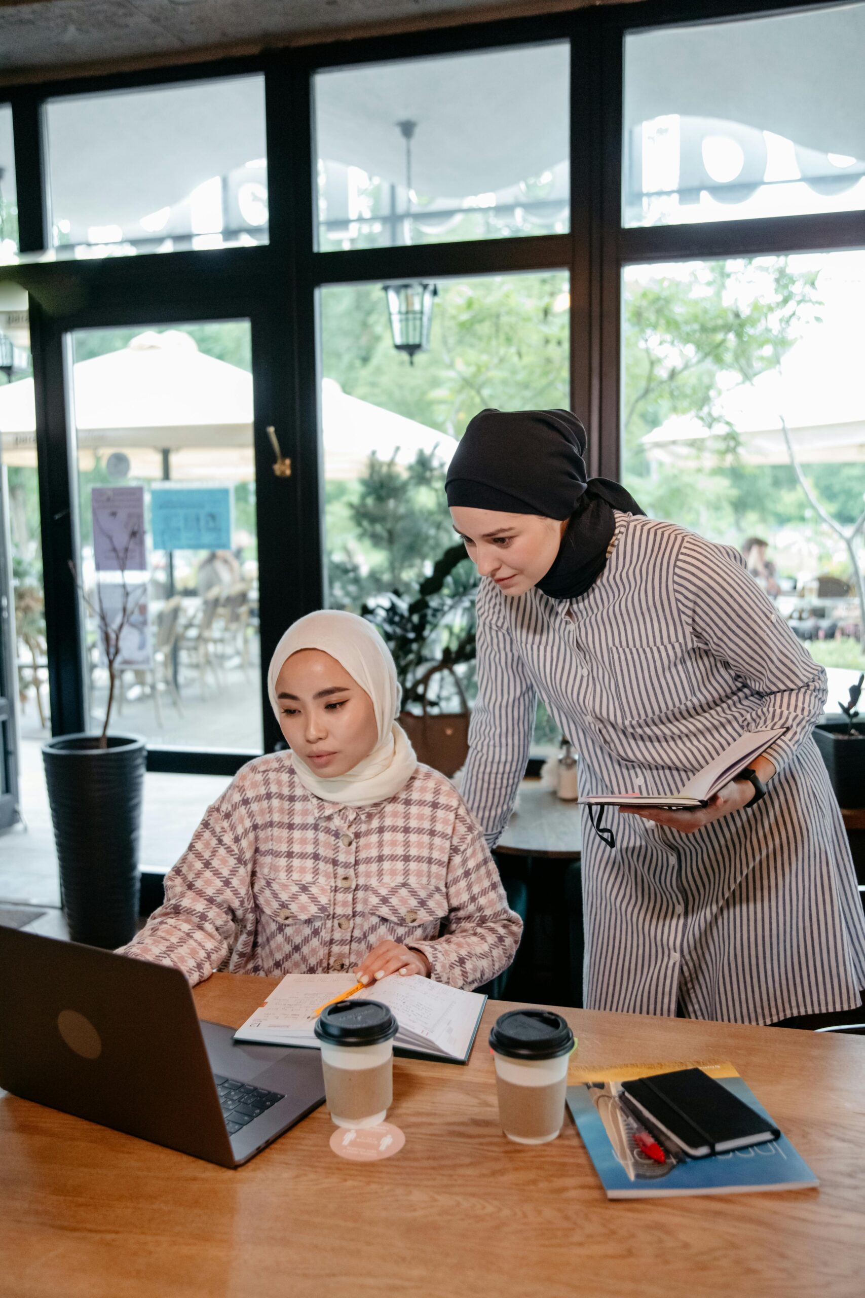 Two Muslim women wearing hijabs collaborate on a project using a laptop in a modern café.